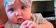 Iris is registered to the contest to win money with this photo: adult_hand, baby, blue_eyes, bow, closeup, clothing, curtain, cute, expression, face, hands, headband, indoor, infant, person, plant, portrait, skin, television, window