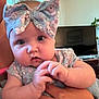 Iris is registered to the contest to win money with this photo: baby, infant, headband, bow, hands, person, face, blue_eyes, clothing, adult_hand, indoor, television, plant, curtain, window, skin, expression, cute, closeup, portrait