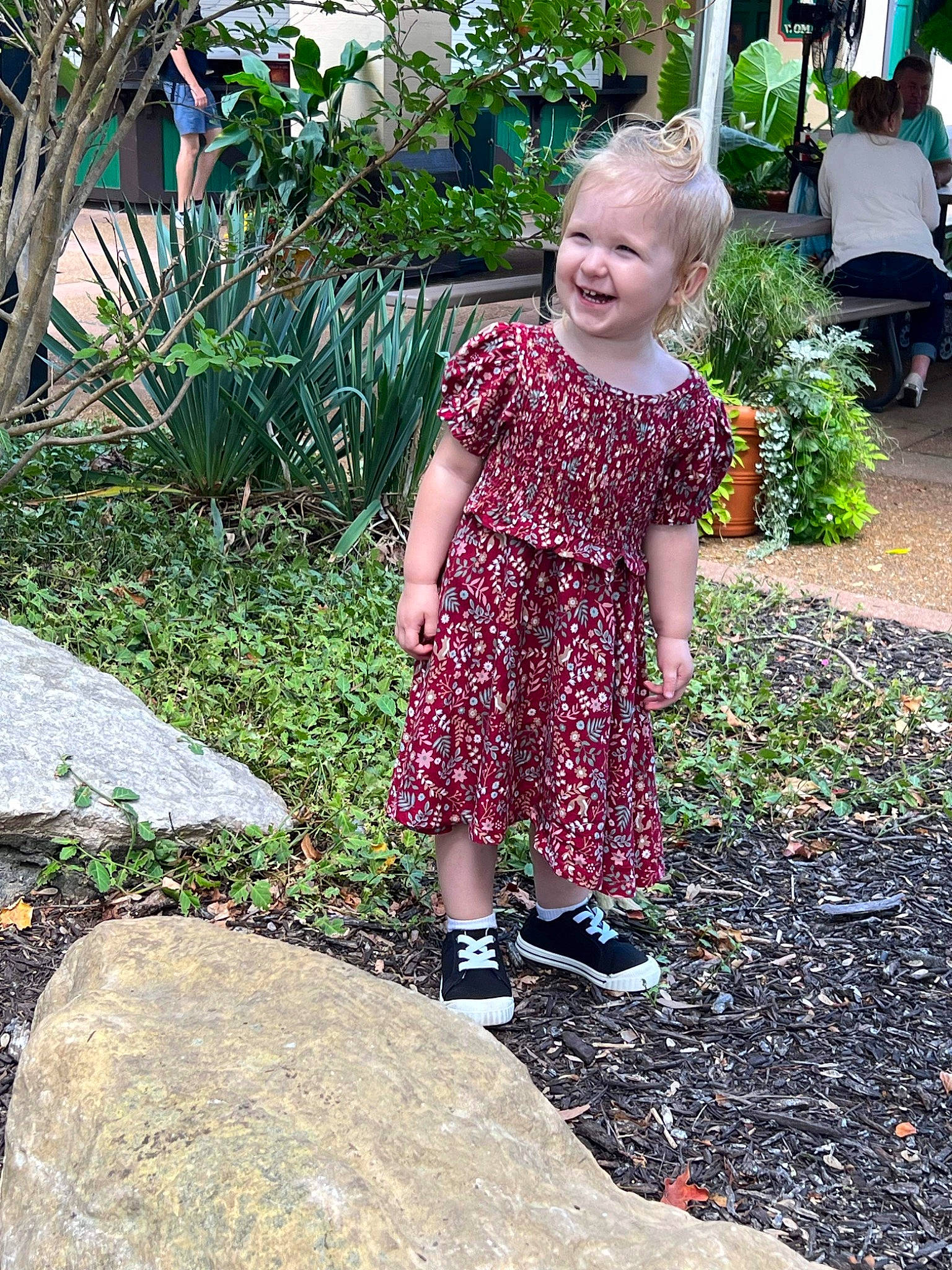 Amelia is registered to the contest to win money with this photo: baby_toddler_clothing, botany, child, dress, fun, garden, grass, green, groundcover, hairstyle, joy, leisure, magenta, pattern, people_in_nature, person, plant, shrub, sitting, smile