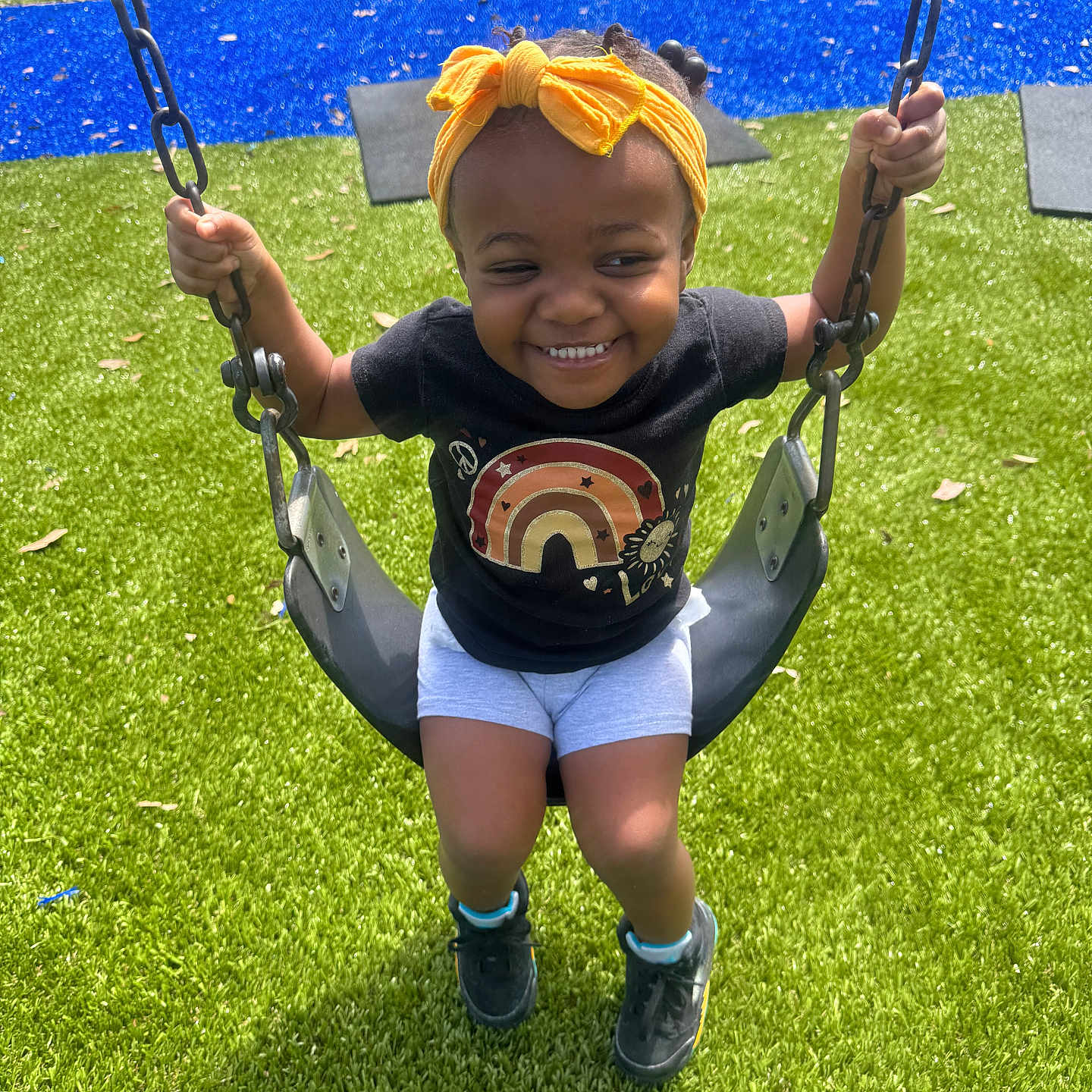 Jahzari is registered to the contest to win money with this photo: child, toddler, swing, playground, grass, artificial_grass, headband, yellow, black_tshirt, rainbow, shoes, happy, smiling, outdoor, play, sunny, daylight, person, young_child, recreation