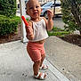 toddler, child, popsicle, sidewalk, concrete, white_top, coral_shorts, white_sandals, bushes, car, suburban, outdoor, summer, playful, expression, person, standing, clothing, daytime, nature
