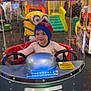 child, beanie, arcade, kiddie_ride, minion, toy, indoor, play_area, steering_wheel, smiling, colorful, amusement, game, people, background, fun, metallic, seat, person, hat
