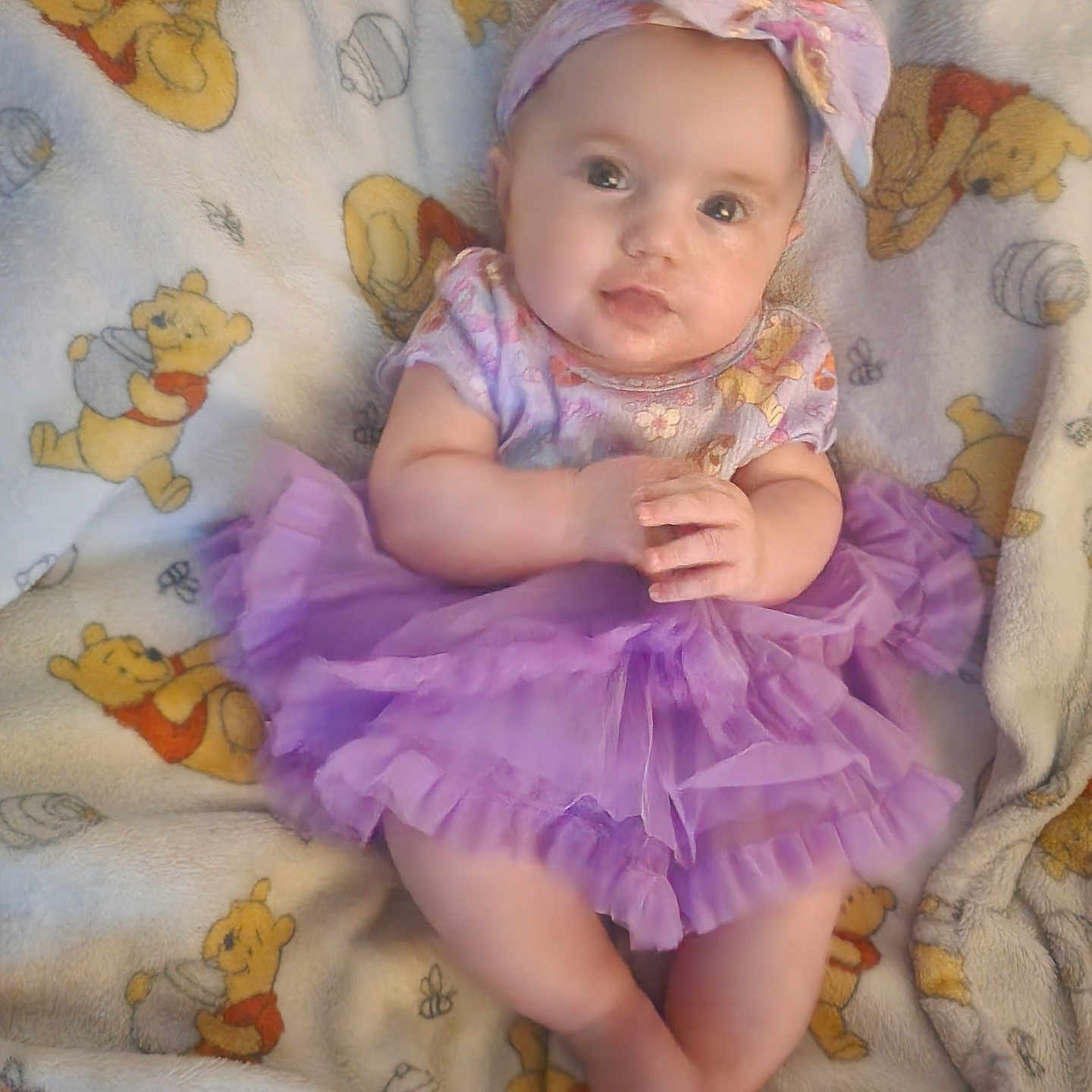 Hennessi is registered to the contest to win money with this photo: baby, infant, child, purple_tutu, headband, blanket, winnie_the_pooh, cute, smiling, portrait, soft_toys, plush_blanket, indoors, sitting, clothing, hands, legs, face, expression, colorful