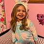 child, unicorn_onesie, smiling, indoor, pink_wall, height_chart, toy_pig, chevron_pattern, bed, furniture, happy, portrait, cute, costume, long_hair, young_child, face, clothing, room, playful