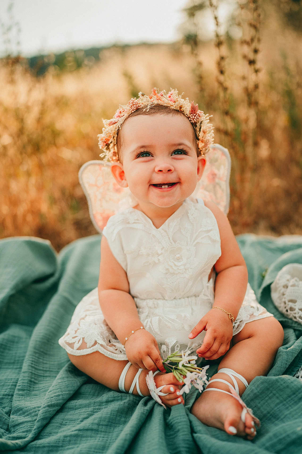 Millie joined the competition — help win amazing prizes! baby, smiling, child, flower_crown, white_dress, lace, outdoor, green_blanket, grass, nature, cute, happy, portrait, toes, flower, sunlight, infant, sitting, bracelet, headband