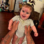 Everleigh joined the competition — help win amazing prizes! baby, chair, child, clothing, door, face, hair_tie, hands, happy, holding, home, indoor, infant, person, playful, smiling, standing, tattoos, wall_decor, wooden_floor