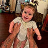 baby, chair, child, clothing, door, face, hair_tie, hands, happy, holding, home, indoor, infant, person, playful, smiling, standing, tattoos, wall_decor, wooden_floor