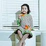 child, girl, barefoot, sitting, stool, window, blinds, indoor, dress, patterned_clothing, holding, hair_clip, calm, contemplative, modern_furniture, floor, portrait, person, home, young_child