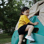 Cing is registered to the contest to win money with this photo: child, toddler, climbing, playground, outdoor, yellow_shirt, black_shorts, green_play_structure, park, nature, trees, shoes, active, play, young_child, summer, daytime, recreation, fun, casual
