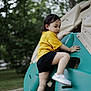 child, toddler, climbing, playground, outdoor, yellow_shirt, black_shorts, green_play_structure, park, nature, trees, shoes, active, play, young_child, summer, daytime, recreation, fun, casual