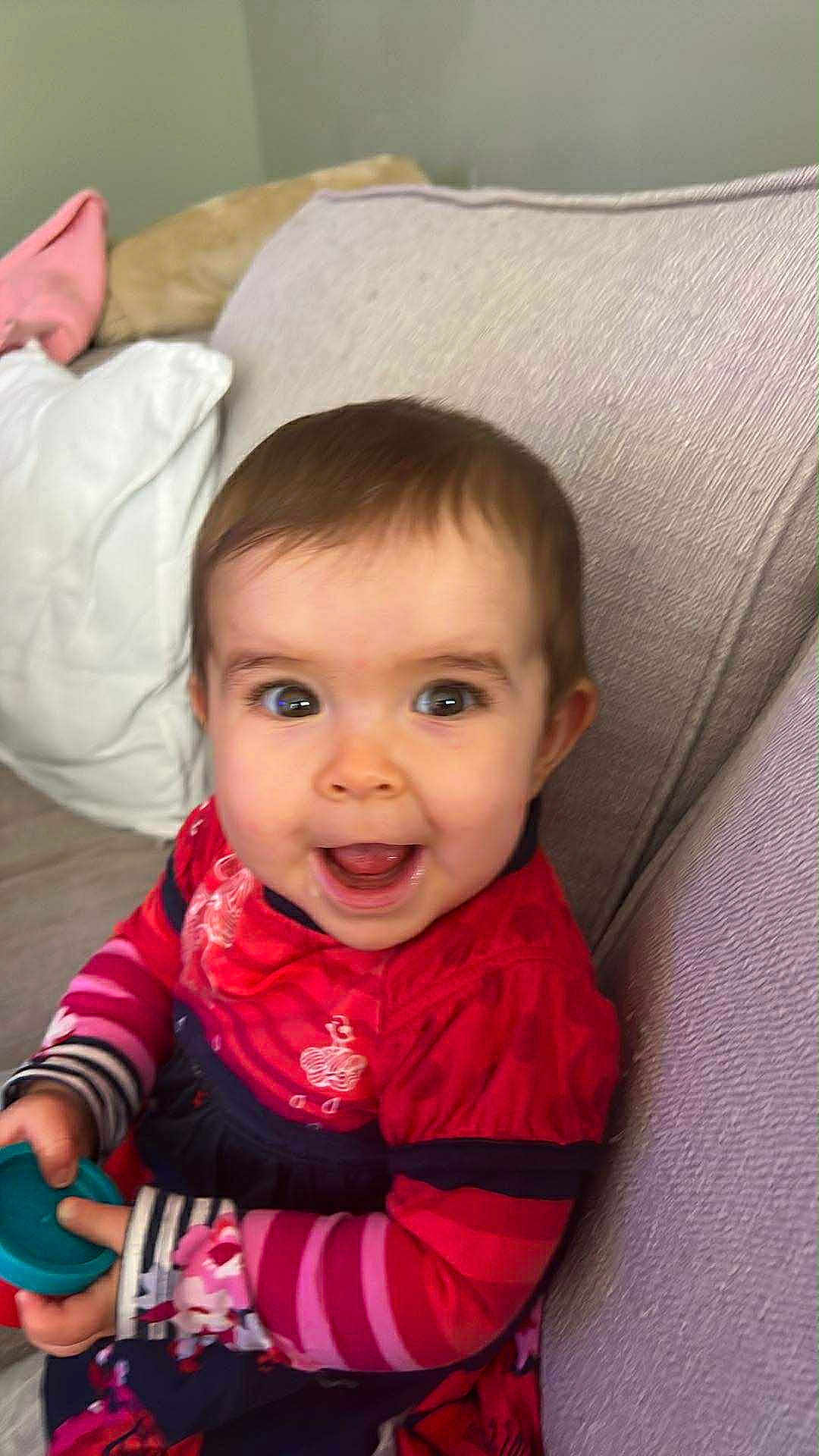 Emy a rejoint le concours — aidez-le/la à gagner de superbes lots ! baby, child, smiling, happy, indoor, couch, toy, clothing, striped_sleeves, person, face, young_child, seated, home, cute, bright_colors, playful, infant, apparel, joyful