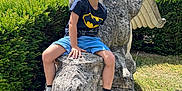 Lilio a rejoint le concours — aidez-le/la à gagner de superbes lots ! child, boy, smiling, outdoor, statue, griffin, stone, garden, greenery, tree, sunlight, batman_tshirt, red_shoes, shorts, playful, happy, nature, portrait, casual, daytime