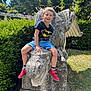 child, boy, smiling, outdoor, statue, griffin, stone, garden, greenery, tree, sunlight, batman_tshirt, red_shoes, shorts, playful, happy, nature, portrait, casual, daytime