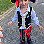 child, pirate_costume, hat, vest, pants, outdoor, grass, pavement, smile, playful, costume, footwear, sidewalk, skull_and_crossbones, red, black, white_shirt, gold_trim, person, fun