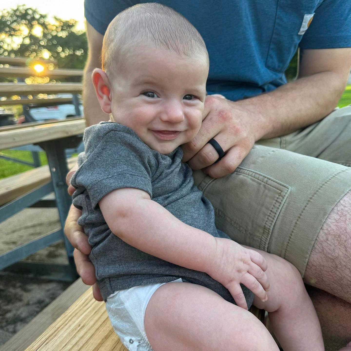 Liam joined the competition — help win amazing prizes! adult, baby, bench, clothing, face, hand, happy, infant, nature, outdoor, park, person, portrait, shorts, skin, smiling, sunlight, trees, tshirt, wood