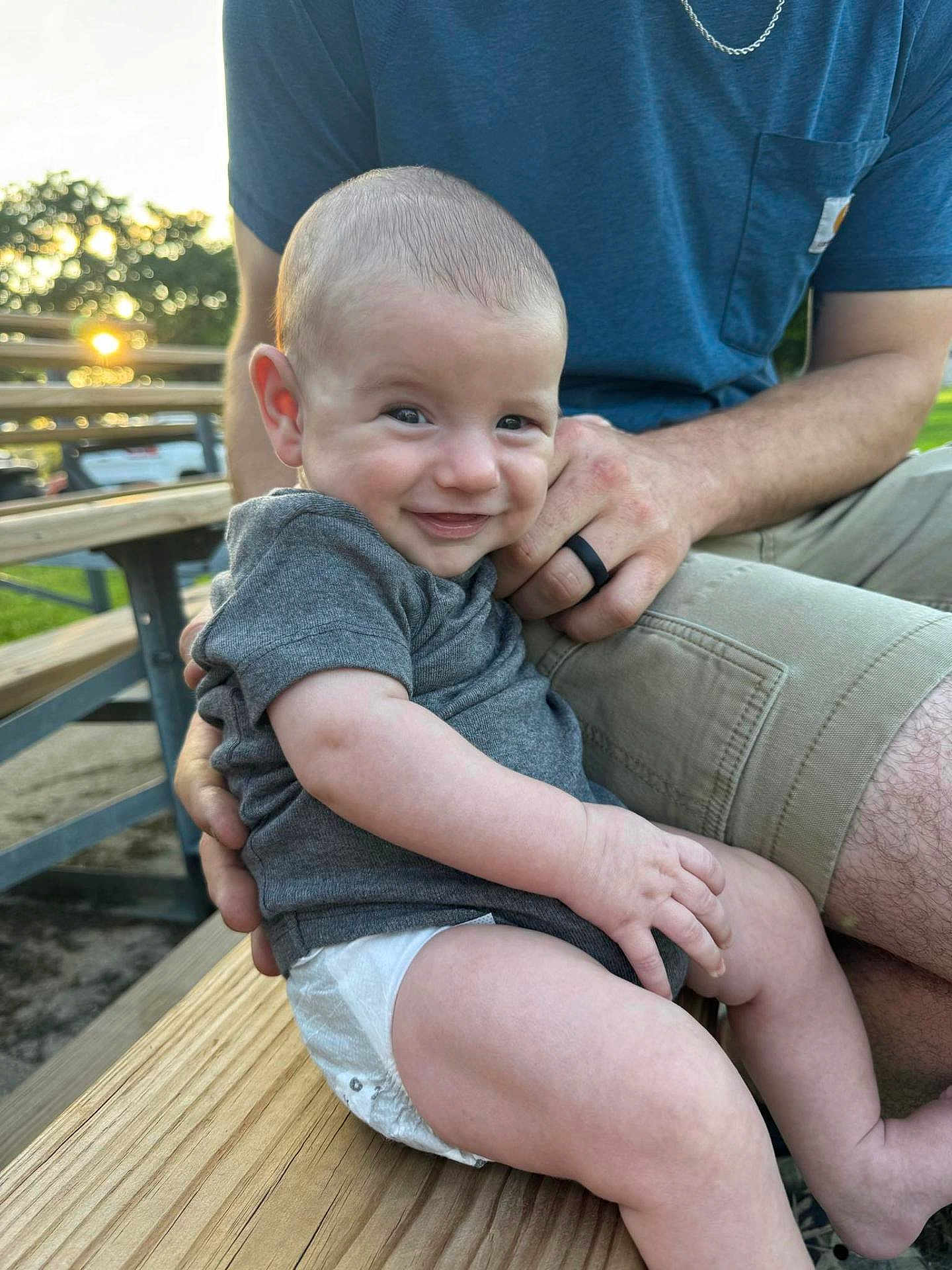 Liam joined the competition — help win amazing prizes! baby, smiling, infant, outdoor, bench, wood, person, adult, hand, clothing, shorts, tshirt, sunlight, nature, trees, park, skin, face, happy, portrait