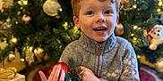 Oliver joined the competition — help win amazing prizes! child, red_hair, smiling, gray_sweater, christmas_tree, ornament, pinecone, gift_box, gold_ribbon, holiday, festive, indoor, happy, decorations, present, seasonal, celebration, young_child, sitting, cozy