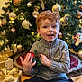 child, red_hair, smiling, gray_sweater, christmas_tree, ornament, pinecone, gift_box, gold_ribbon, holiday, festive, indoor, happy, decorations, present, seasonal, celebration, young_child, sitting, cozy