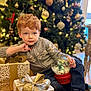 child, red_hair, christmas_tree, gift_box, snow_globe, holiday, festive, decorations, indoor, floor, sweater, jeans, holiday_lighting, present, celebration, winter_clothing, home, relaxed, holiday_season, smiling