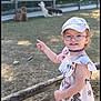 alpaca, animal, cap, child, cute, daytime, dress, fence, girl, glasses, grass, happy, nature, outdoor, pointing, shoes, smiling, socks, sunlight, trees