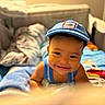 child, toddler, baby, smiling, portrait, face, blue_cap, tank_top, bed, bedroom, blankets, bedding, indoors, cozy, warm_lighting, happy, lying_down, looking_at_camera, home, photography