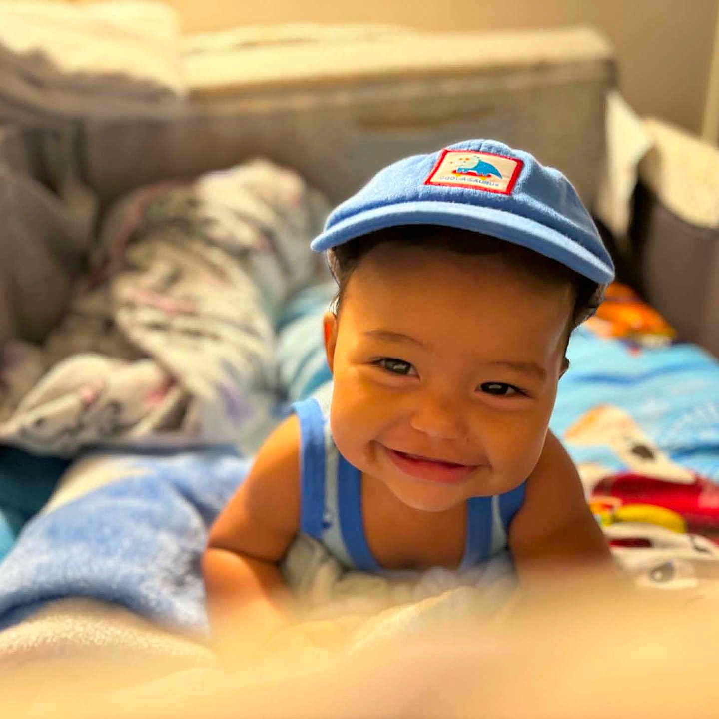 Jaci is registered to the contest to win money with this photo: child, toddler, baby, smiling, portrait, face, blue_cap, tank_top, bed, bedroom, blankets, bedding, indoors, cozy, warm_lighting, happy, lying_down, looking_at_camera, home, photography