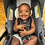 baby, smiling, stroller, child, happy, infant, seatbelt, indoor, wooden_floor, black_clothing, shorts, hands, feet, face, ears, cute, safety_harness, person, portrait, closeup