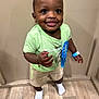 toddler, child, smiling, standing, indoor, green_shirt, shorts, blue_ribbon, bracelet, necklace, socks, flooring, wood_floor, face, happy, cute, person, portrait, young_child, champion