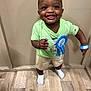 toddler, child, smiling, birthday, blue_ribbon, green_shirt, shorts, socks, floor, wooden_floor, bracelet, necklace, earrings, wristband, indoor, happy, person, standing, celebration, portrait