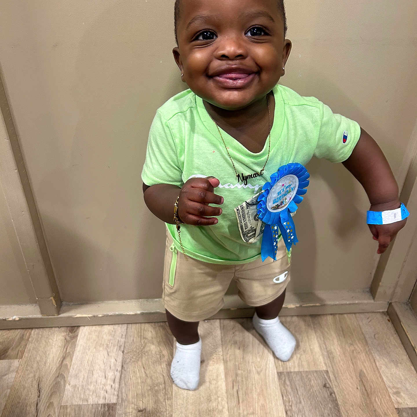 Nymari is registered to the contest to win money with this photo: birthday, blue_ribbon, bracelet, celebration, child, earrings, floor, green_shirt, happy, indoor, necklace, person, portrait, shorts, smiling, socks, standing, toddler, wooden_floor, wristband