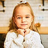 background_blur, blonde_hair, casual_clothing, child, cozy, cute, earrings, face, girl, hair_bow, home, indoors, kitchen, person, portrait, resting_chin, smiling_slightly, warm_lighting, white_sweater, young