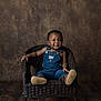 toddler, child, wicker_chair, denim_overalls, brown_boots, jewelry, bracelet, smiling, portrait, studio, brown_background, wooden_floor, sitting, curly_hair, happy, indoor, fashion, cute, young_child, casual_clothing