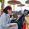 woman, dog, beach, stroller, umbrella, sand, outdoor, sunny, casual_clothing, glasses, smiling, leisure, vacation, lounge_chair, palm_tree, daytime, relaxation, pet, seaside, friendship
