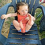 black_chair, blue_eyes, blue_shorts, casual_clothing, child, curious_expression, curly_hair, daylight, feet_up, grass, nature, orange_tank_top, outdoor, person, plastic_chair, playful, sitting, summer, toddler, young_child