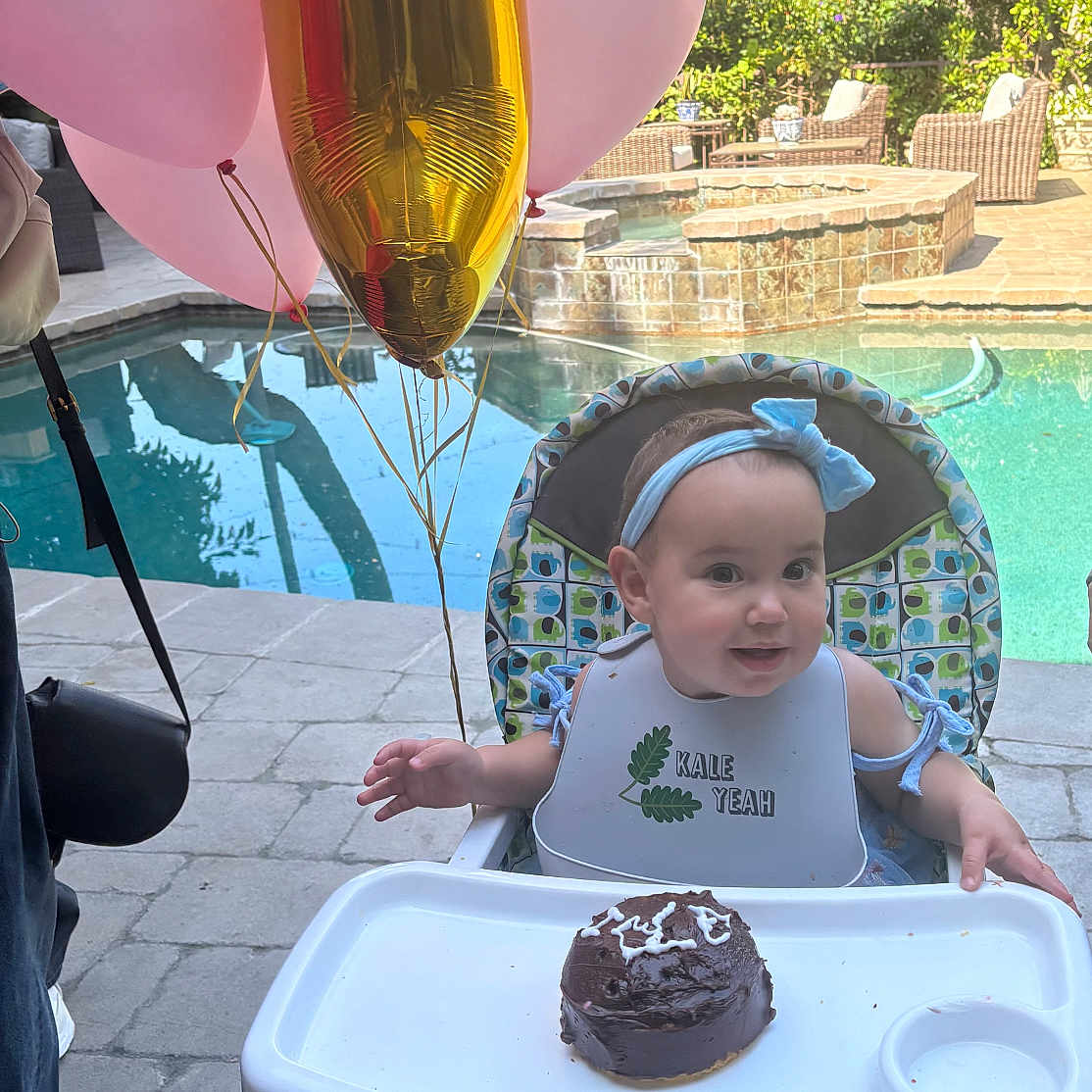 Rebecca joined the competition — help win amazing prizes! baby, high_chair, cake, balloons, pool, outdoor, celebration, headband, bib, smiling, child, chocolate_cake, party, sunlight, footwear, pavement, greenery, wicker_chair, water, fun