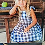 child, girl, smiling, pigtails, dress, blue_and_white, checkered, apple, book, wooden_table, wooden_chair, brick_wall, face, portrait, sitting, indoors, happy, blonde_hair, person, photograph