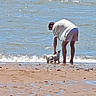 person, dog, beach, shoreline, ocean, water, waves, sand, seaside, outdoor, summer, crouching, pet, animal, white_clothing, shorts, sunlight, coast, travel, beach_walk