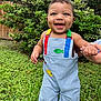 baby, child, blue_eyes, dimples, overalls, colorful_clothing, grass, greenery, outdoor, smiling, happy, person, holding_hand, nature, bushes, fence, daylight, cute, portrait, standing
