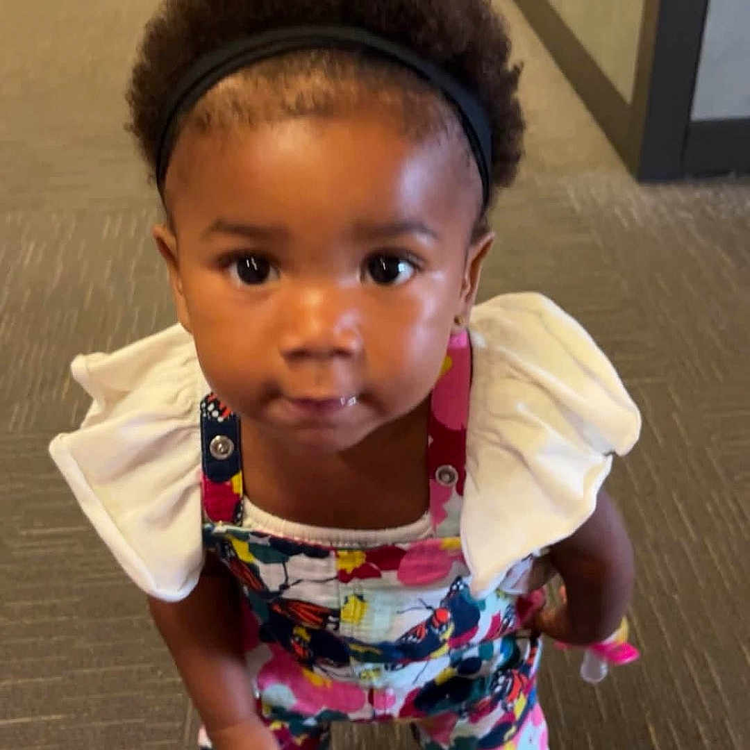 Keore is registered to the contest to win money with this photo: toddler, child, headband, curly_hair, colorful_clothing, standing, indoor, carpet, stroller, baby, cute, looking_up, person, young_child, playful, bright_colors, footwear, small_person, floor, expression