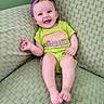Marnie is registered to the contest to win money with this photo: baby, smiling, infant, purple_bow, green_onesie, text_on_clothing, couch, green_background, happy, child, indoor, portrait, cute, sitting, headband, skin, feet, hand, face, person
