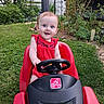 baby, child, red_dress, toy_car, steering_wheel, grass, garden, plants, greenery, outdoor, smile, happy, cute, play, summer, yard, fence, nature, person, portrait