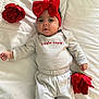 baby, child, red_bow, headband, striped_clothing, flower, red_flower, bed, white_bedspread, lying_down, infant, cute, portrait, face, hands, soft_lighting, indoor, person, smiling, young_child