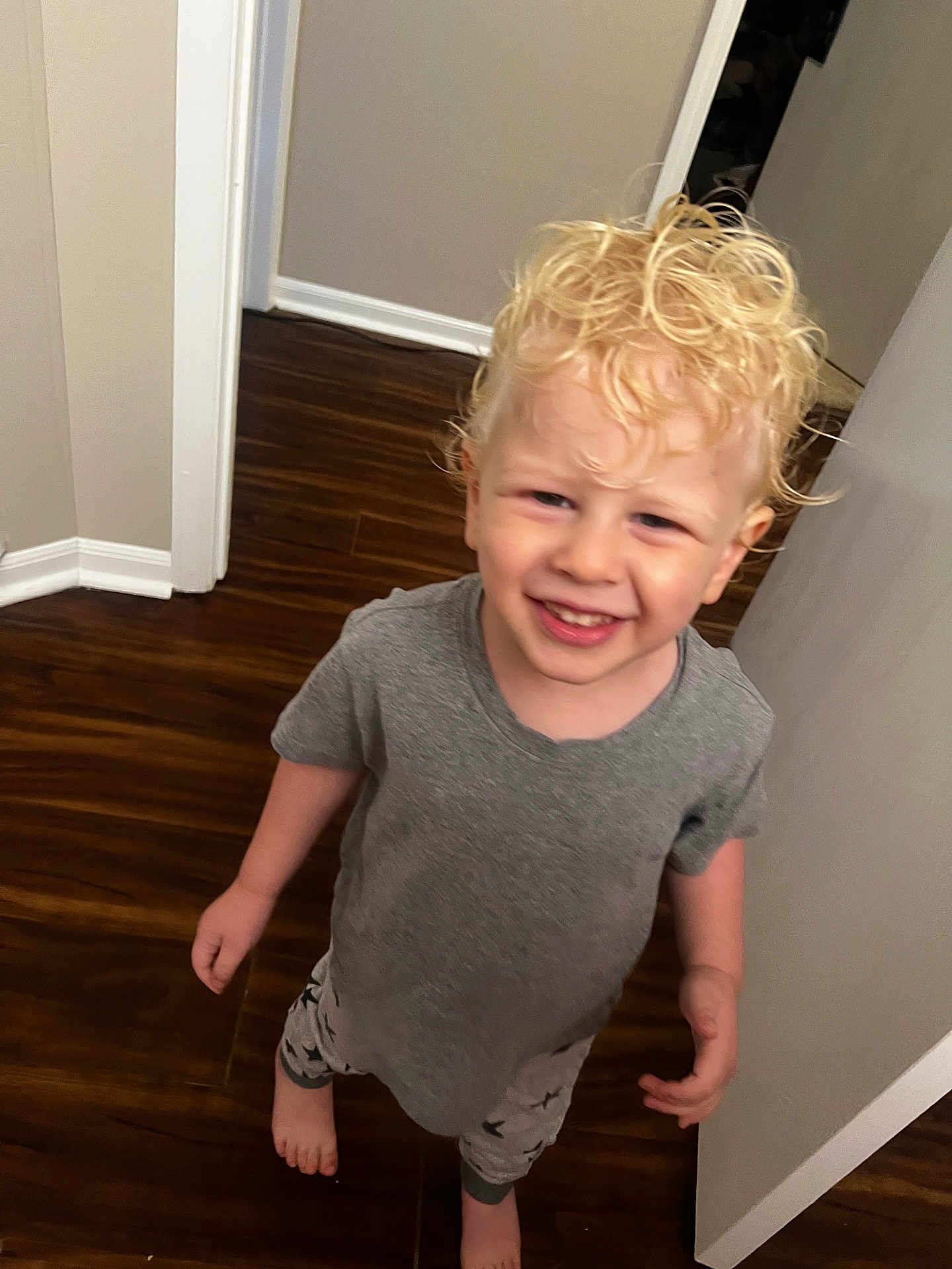 Avryall Apollo is registered to the contest to win money with this photo: child, toddler, smiling, curly_hair, blonde, grey_tshirt, star_patterned_pants, barefoot, wooden_floor, indoor, hallway, doorway, happy, person, young_child, casual_clothing, standing, home, bright, portrait