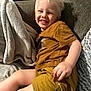 child, smiling, blonde_hair, shorts, tshirt, couch, blanket, indoor, happy, relaxed, cozy, person, young_child, portrait, casual_clothing, furniture, home, comfort, cute, sitting