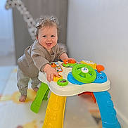 Elio a rejoint le concours — aidez-le/la à gagner de superbes lots ! baby, infant, smiling, play_table, activity_center, colorful_toy, standing, hands, face, hair, grey_sweater, grey_pants, barefoot, nursery, playmat, indoor, joyful, steering_wheel, orange_button, plastic_legs