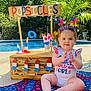 baby, child, popsicles, pool, summer, patriotic, red_white_blue, blanket, outdoor, sunny, headband, clapping, sign, wooden_stand, plants, water, fun, celebration, happy, person
