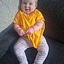 baby, child, smile, yellow_shirt, floral_pants, barefoot, couch, indoor, happy, sitting, face, person, cute, infant, plump, clothing, portrait, cozy, home, skin