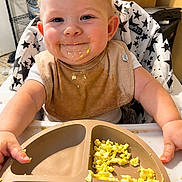 Jermaine is registered to the contest to win money with this photo: baby, child, high_chair, messy_face, bib, scrambled_eggs, food, plate, smiling, happy, indoor, meal_time, toddler, cute, face, hands, eating, kitchen, food_mess, infant