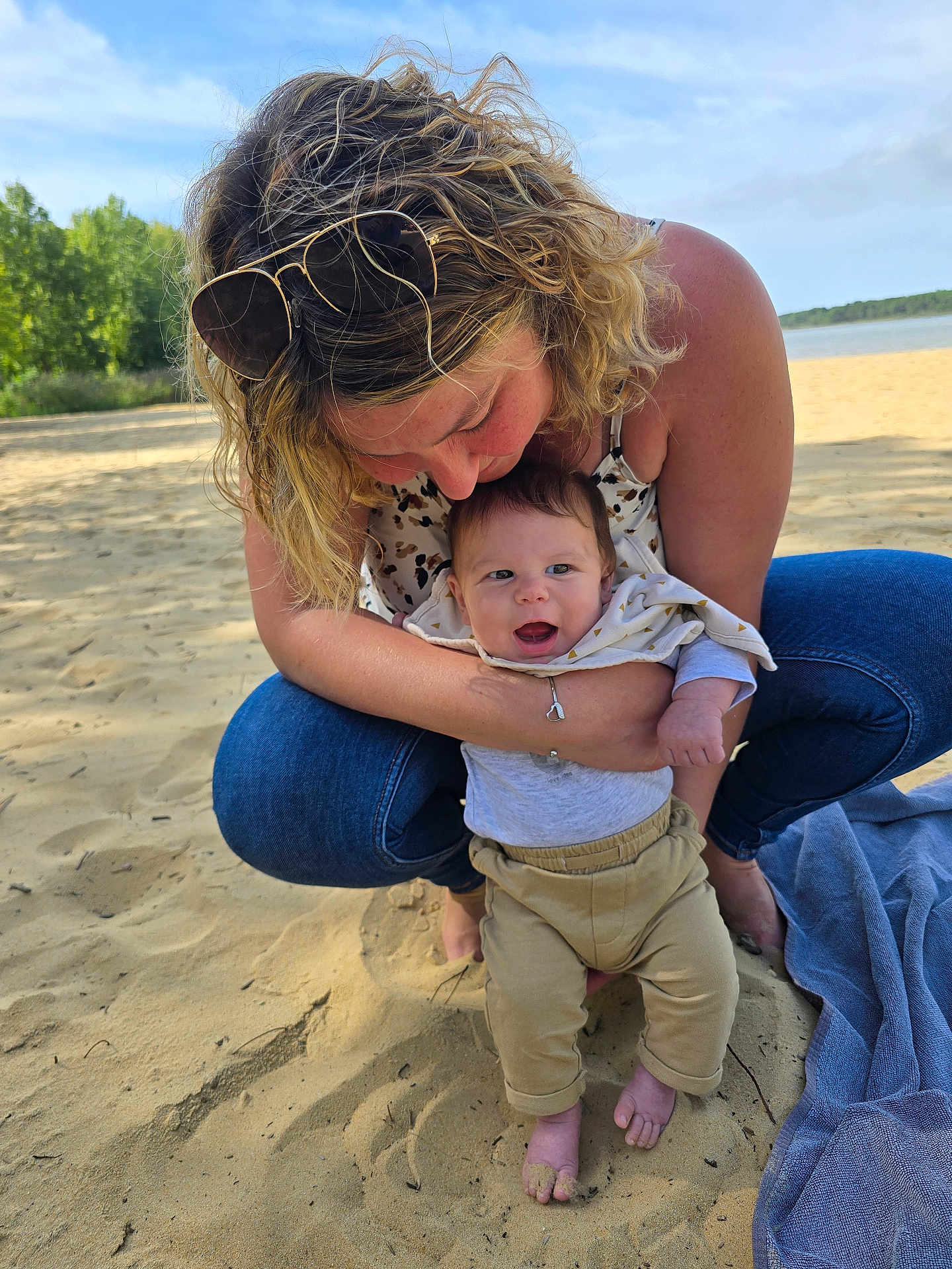 Nolann a rejoint le concours — aidez-le/la à gagner de superbes lots ! baby, woman, beach, sand, water, trees, sunlight, smile, happy, casual_clothing, jeans, sunglasses, blanket, nature, outdoor, family, affection, child, portrait, barefoot