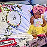 baby, child, pink_bow, yellow_tutu, floral_blanket, numbers, leafy_wreath, sitting, cute, indoor, portrait, clothing, infant, soft_texture, decorative, colorful, adorable, face, expression, young